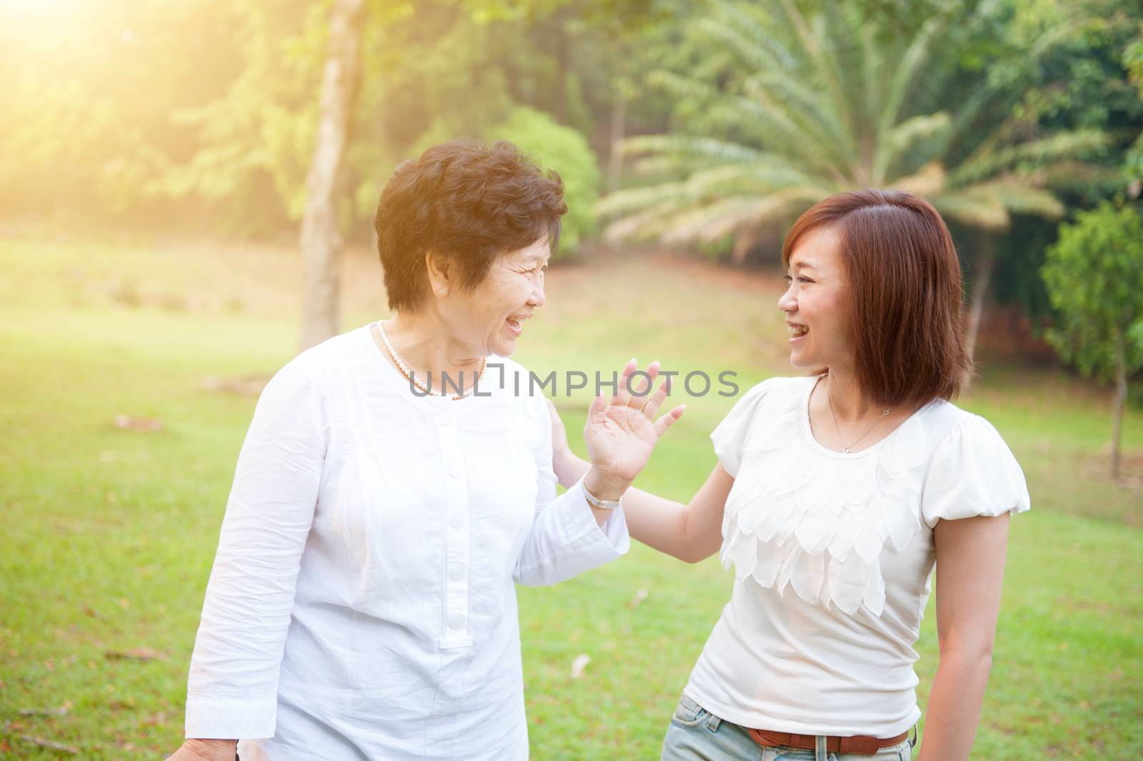 Portrait of happy Asian elderly mother and daughter talking, senior adult woman and grown child. Outdoors family at nature park with beautiful sun flare.