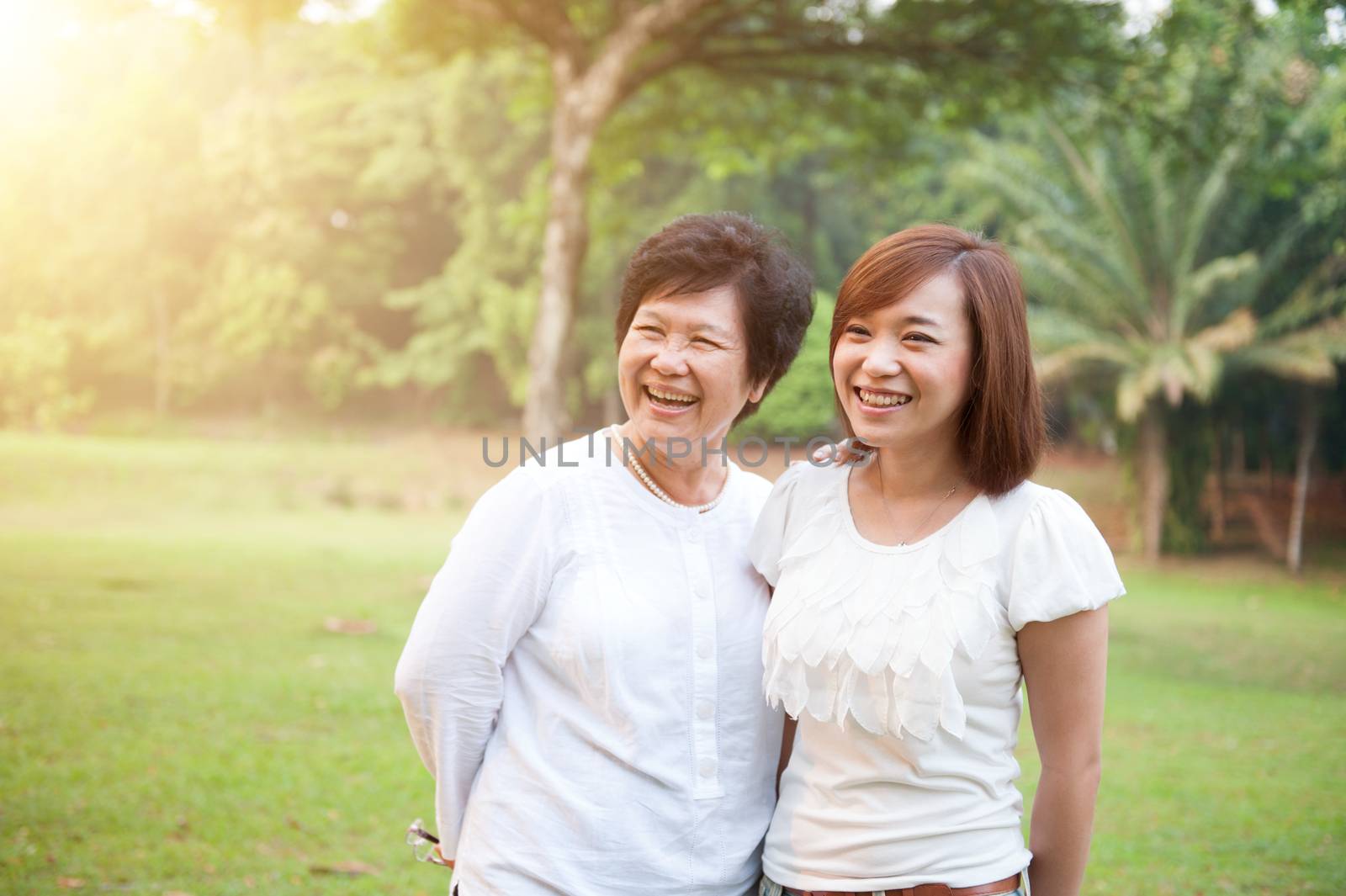 Portrait of happy Asian elderly mother and daughter, senior adult woman and grown child. Outdoors family at nature park with beautiful sun flare.