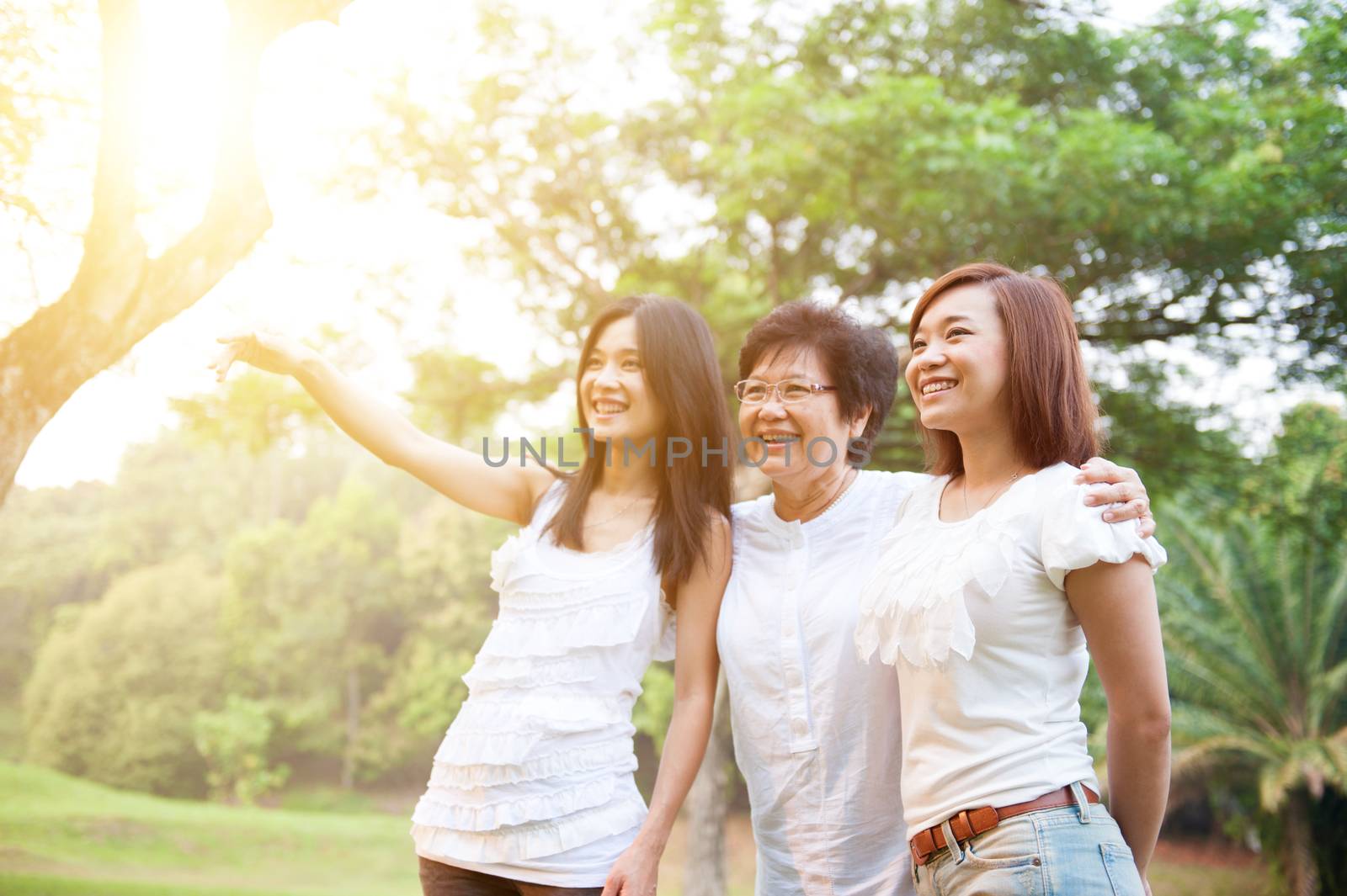 Portrait of Asian elderly mother and daughters pointing looking away, senior adult woman and grown child. Outdoors family at nature park with beautiful sun flare.