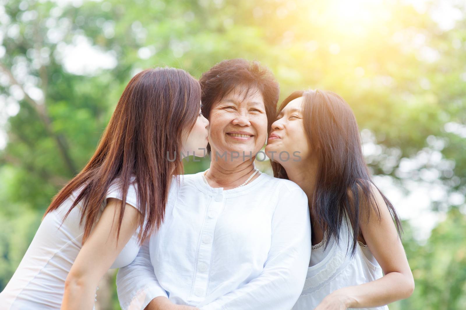 Portrait of Asian daughters kissing elderly mother, senior adult woman and grown child. Outdoors family at nature park with beautiful sun flare.