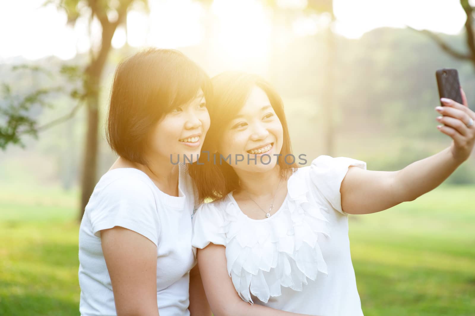 Young Asian females having fun at outdoor park, taking selfie using mobile phone camera, sun flare background.