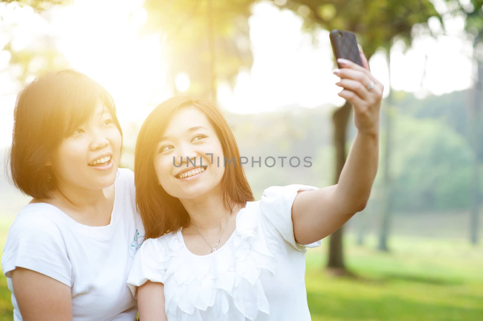 Young Asian females having fun at outdoor park, taking selfie using smartphone camera, sun flare background.