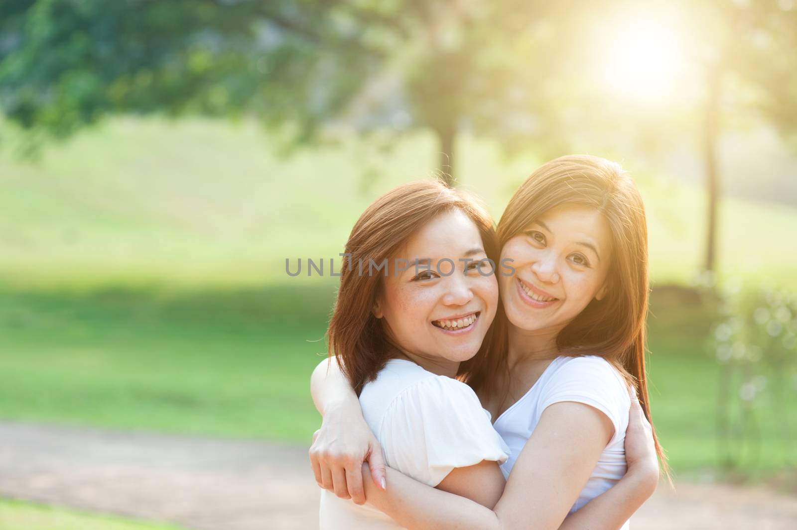 Young Asian sisters having fun at outdoor park, looking at camera and smiling, friendship concept, sun flare background.