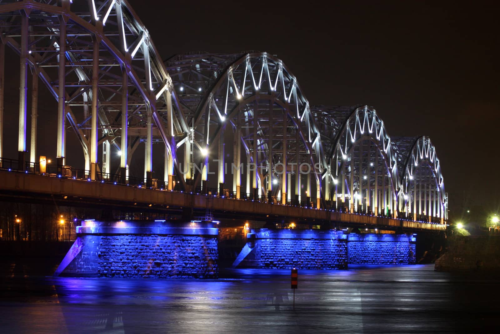 Railway bridge at night with white-blue illumination, reflection in the river