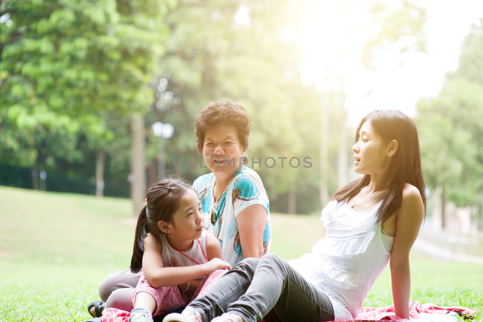 Candid portrait of multi generation Asian family at nature park. Grandmother, mother and grandchild outdoor fun. Morning sun flare background.
