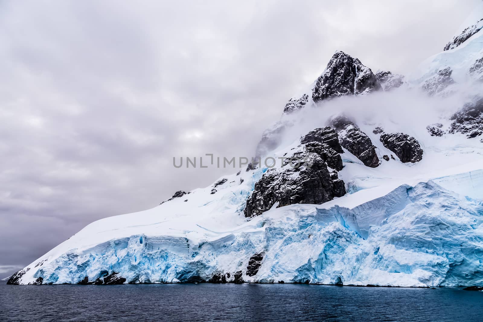 Huge steep stone rock covered with blue glacier and cloud with water in foreground, close to Argentine islands, Antarctica cruise