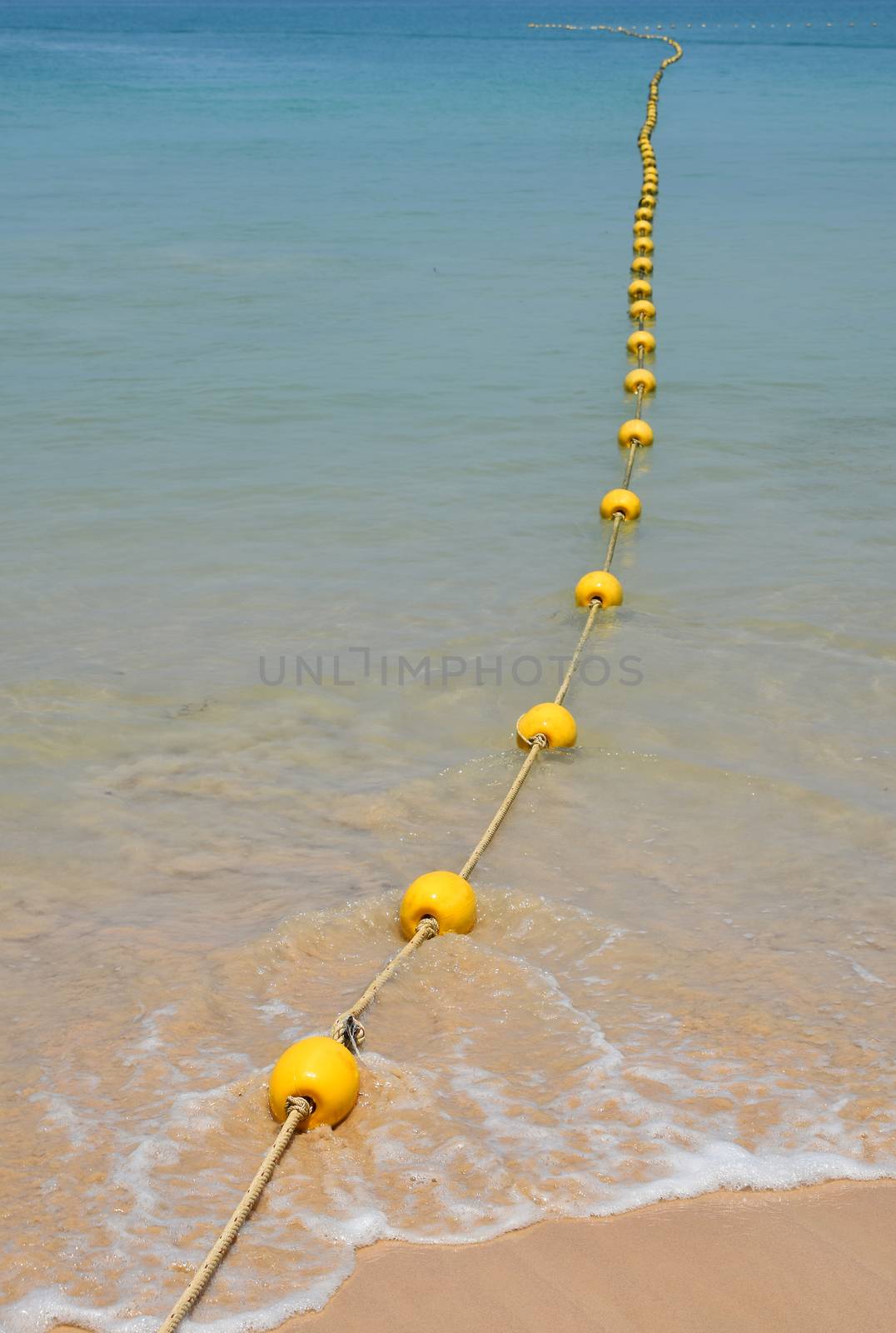 Chain of yellow polystyrene sea marker buoys with cable tow at sand beach and in blue sea water, perspective