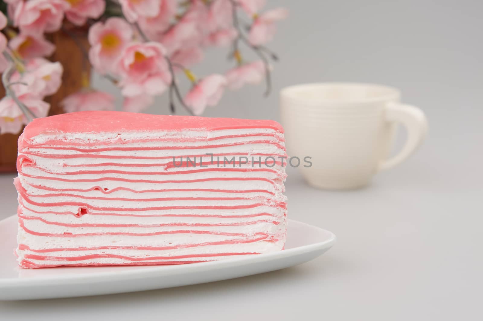Piece of layer cream cake have blur cup of coffee and pink flower as background on a white plate at table.