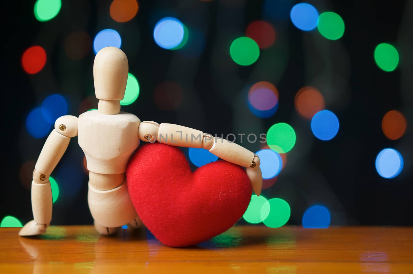 Wooden dummy hug red heart shape and sit on wooden table have colorful bokeh as background.