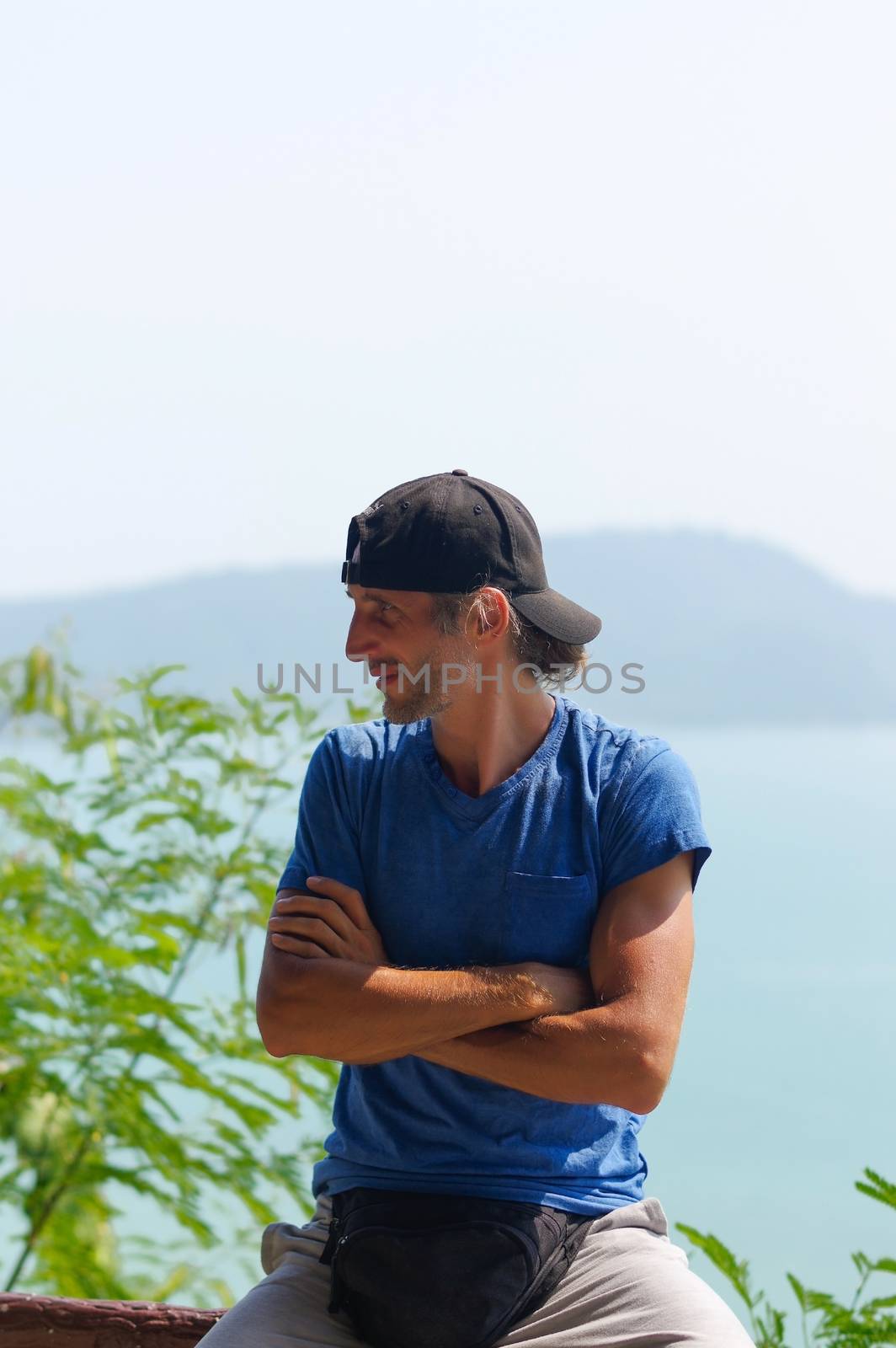 a happy Young Bearded Man sitting on the stone railings in city park wearing in blank blue t-shirt and black Cap