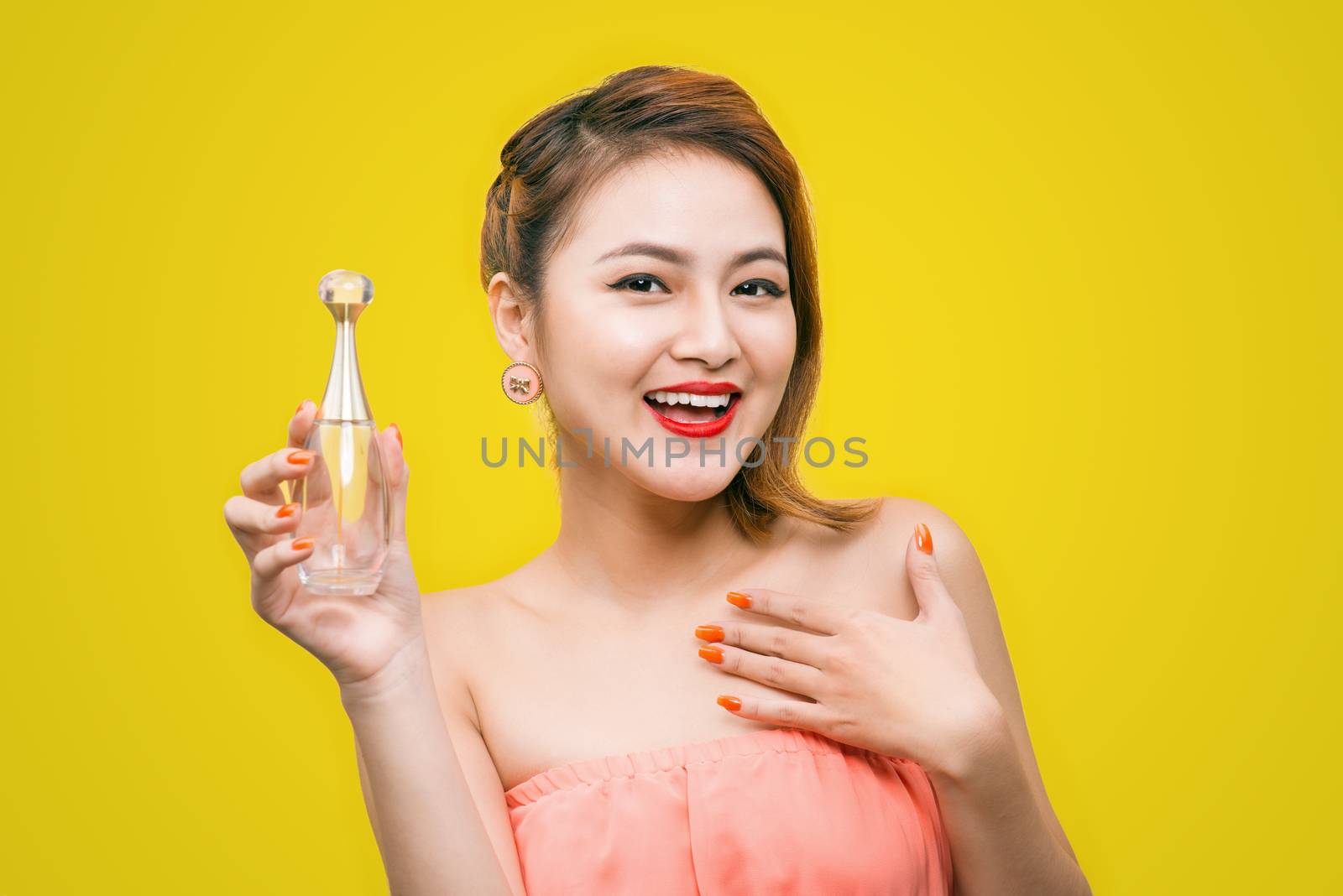 Cheerful beautiful asian young woman with charming smile holding perfume over yellow background.