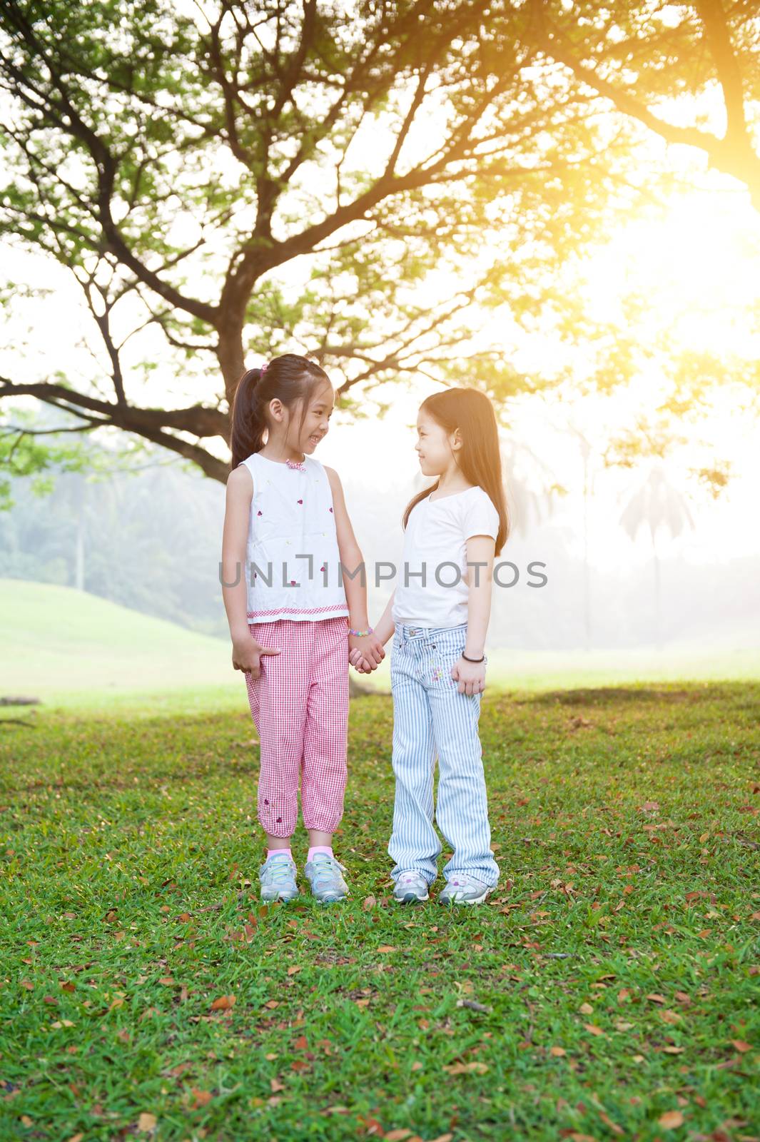 Portrait of Asian children holding hands at park. Little girls having fun outdoors. Morning sun flare background.