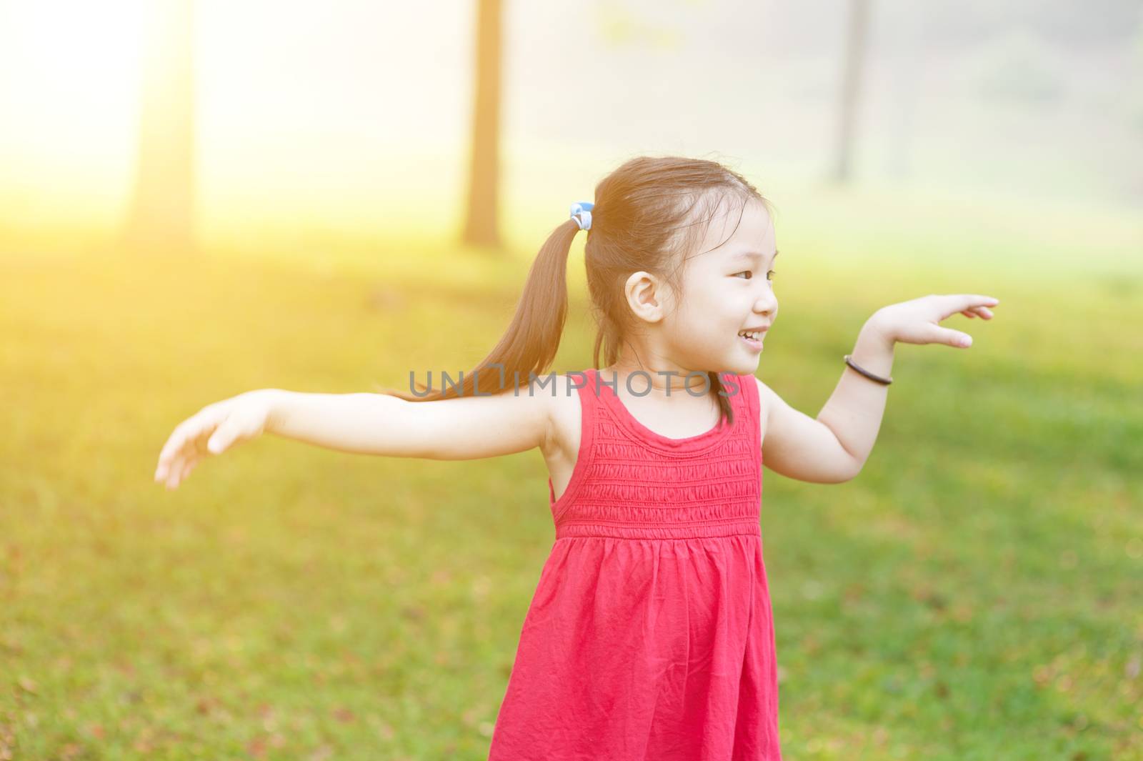 Portrait of active Asian child dancing outdoors. Little girl having fun at nature park. Morning sun flare background.