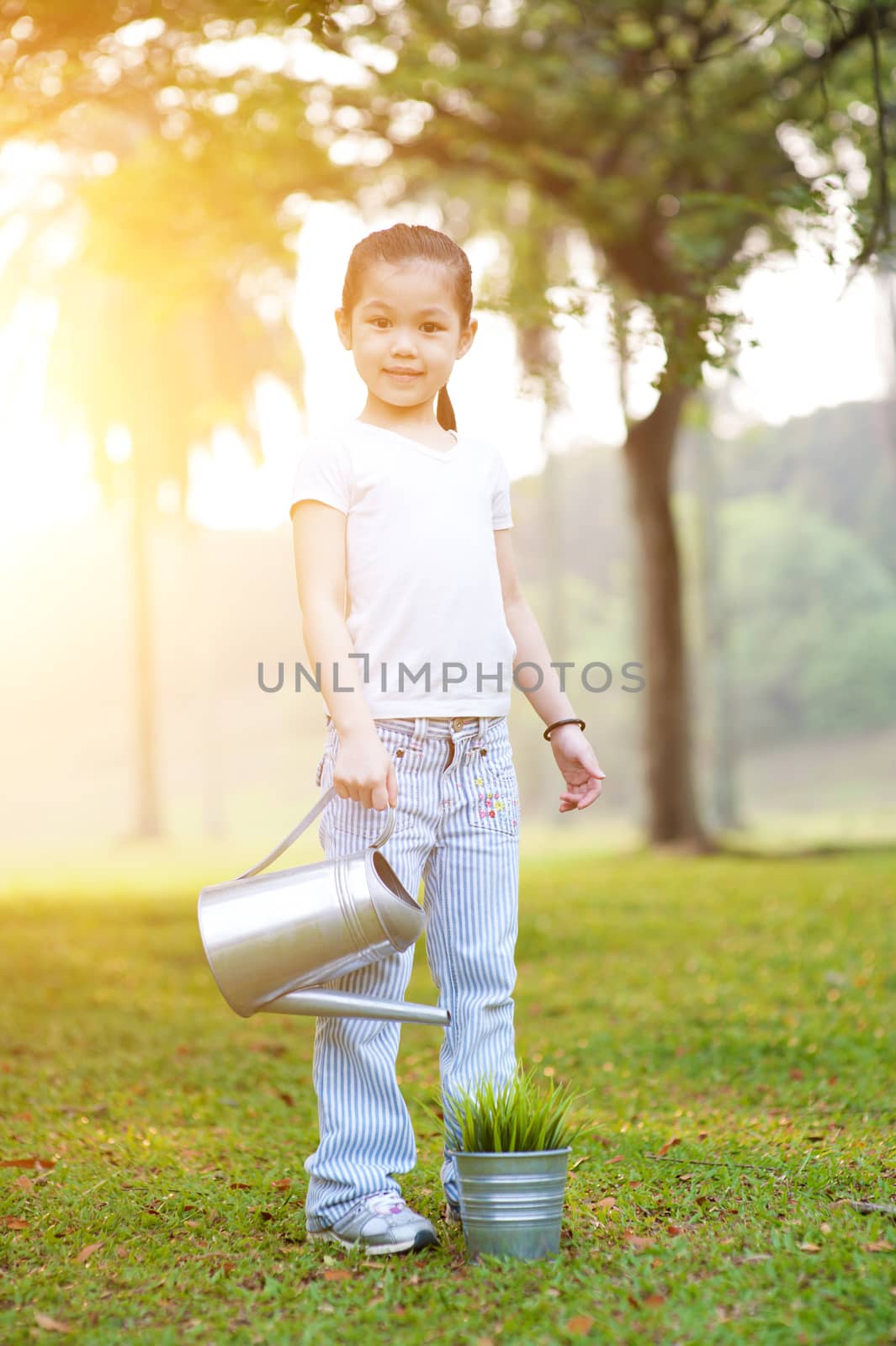 Portrait of active Asian kid watering plant outdoors. Little girl having fun at nature park. Morning sun flare background.