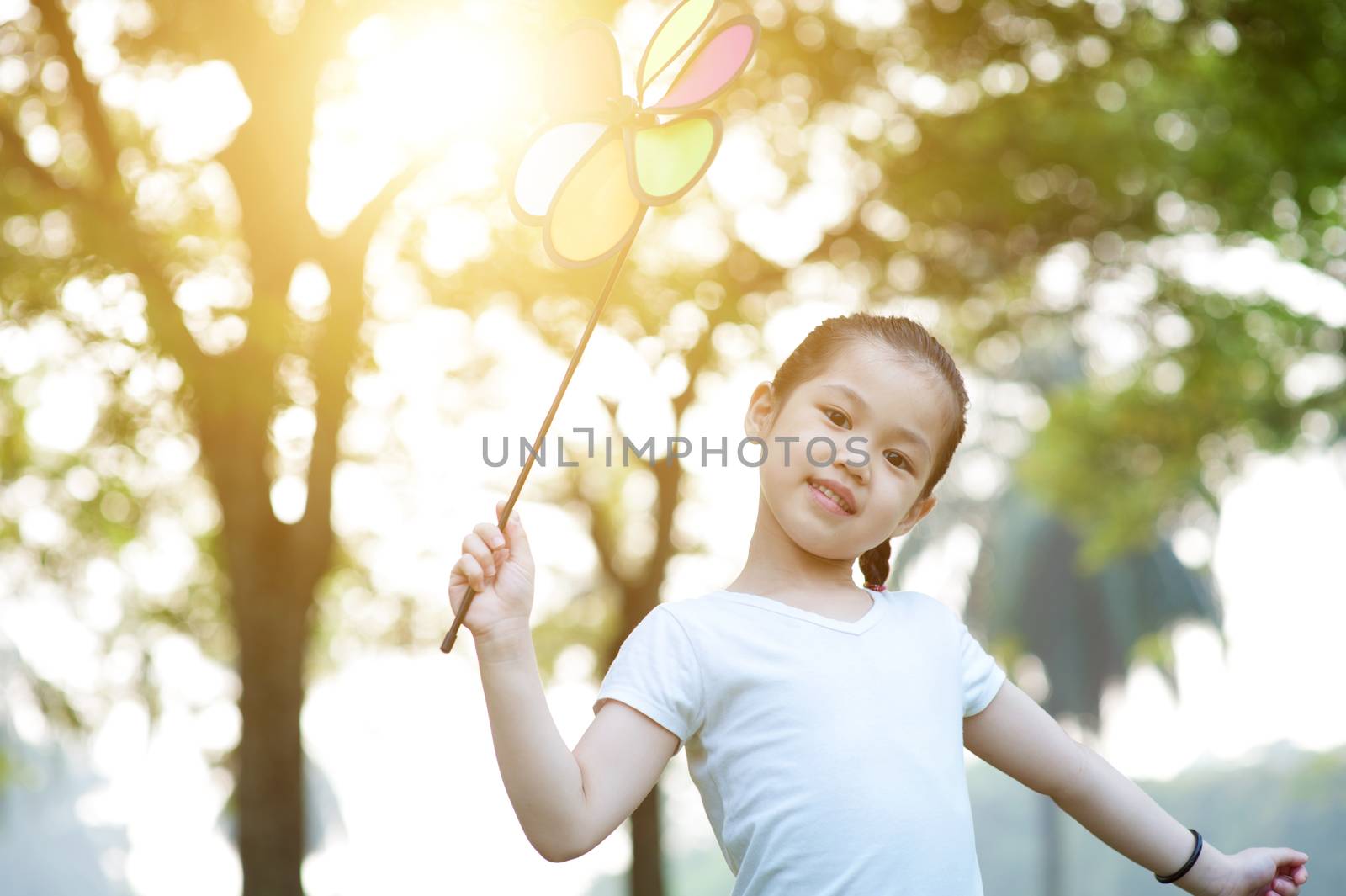 Portrait of cute Asian child playing pinwheel toy outdoors. Little girl having fun at nature park. Morning sun flare background.