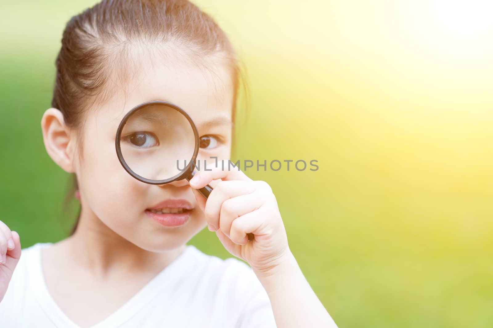 Portrait of cute Asian kid with magnifier glass exploring nature at park. Little girl having fun outdoors. Morning sun flare background.