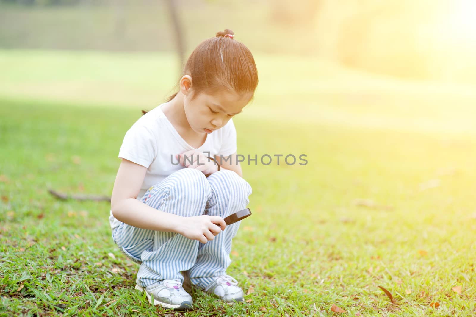 Portrait of Asian school kid with magnifier glass exploring nature at park. Little girl having fun outdoors. Morning sun flare background.