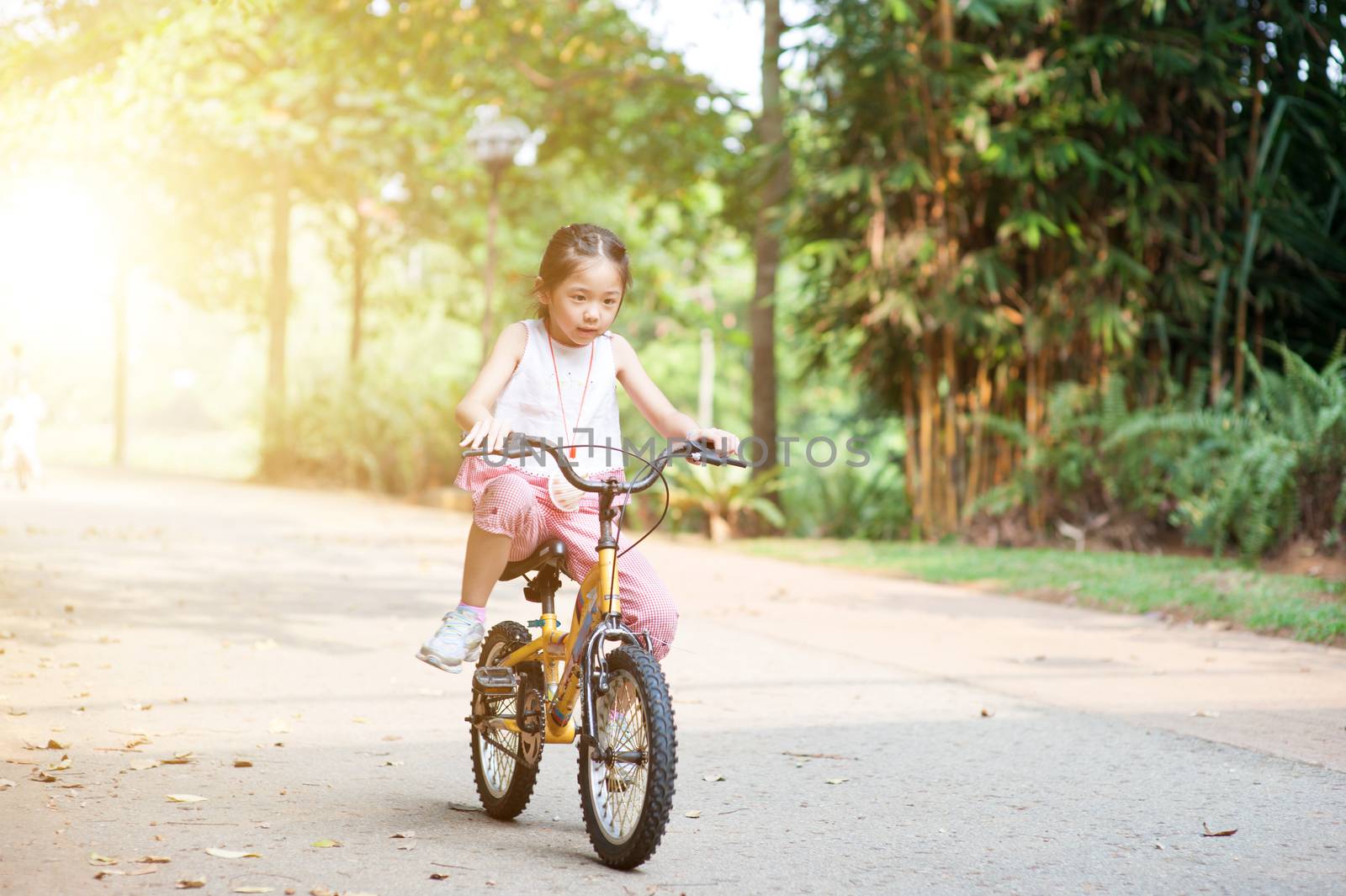 Portrait of active Asian child riding bike outdoors. Kid having fun at nature park. Morning sun flare background.