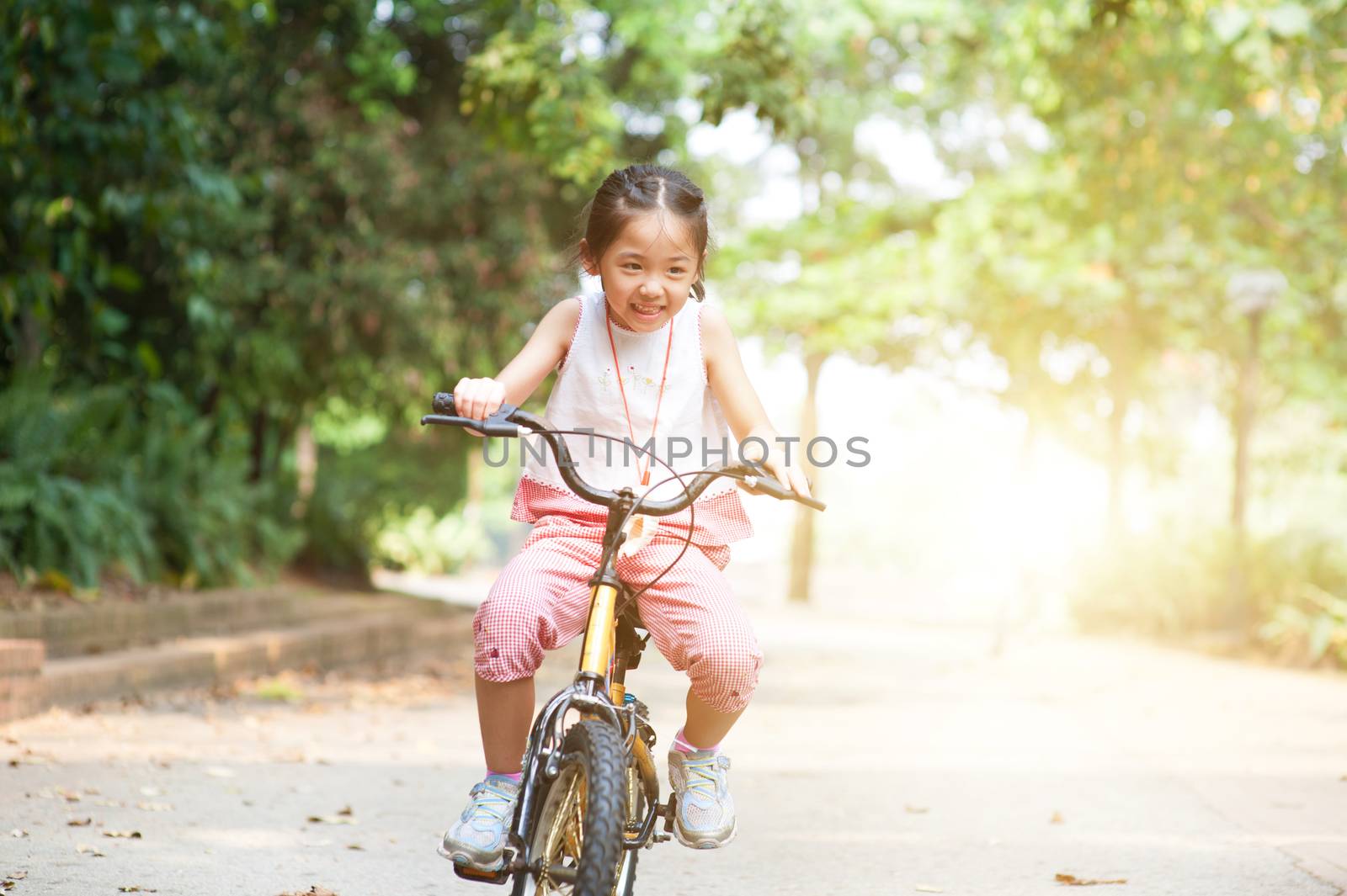 Portrait of active Asian kid riding bicycle outdoors. Little girl having fun at nature park. Morning sun flare background.