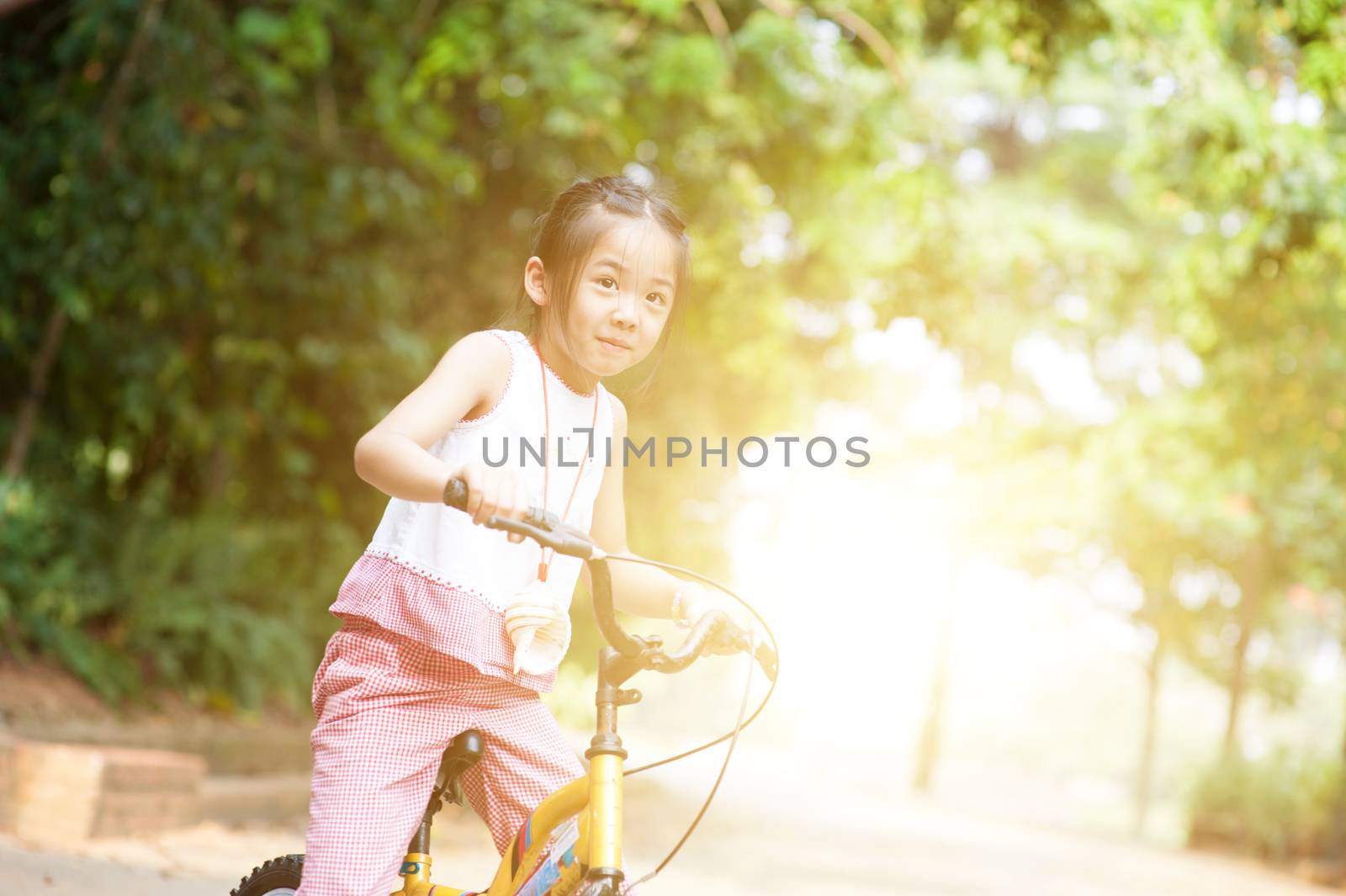 Portrait of active Asian child riding bicycle outdoors. Little girl having fun at nature park. Morning sun flare background.