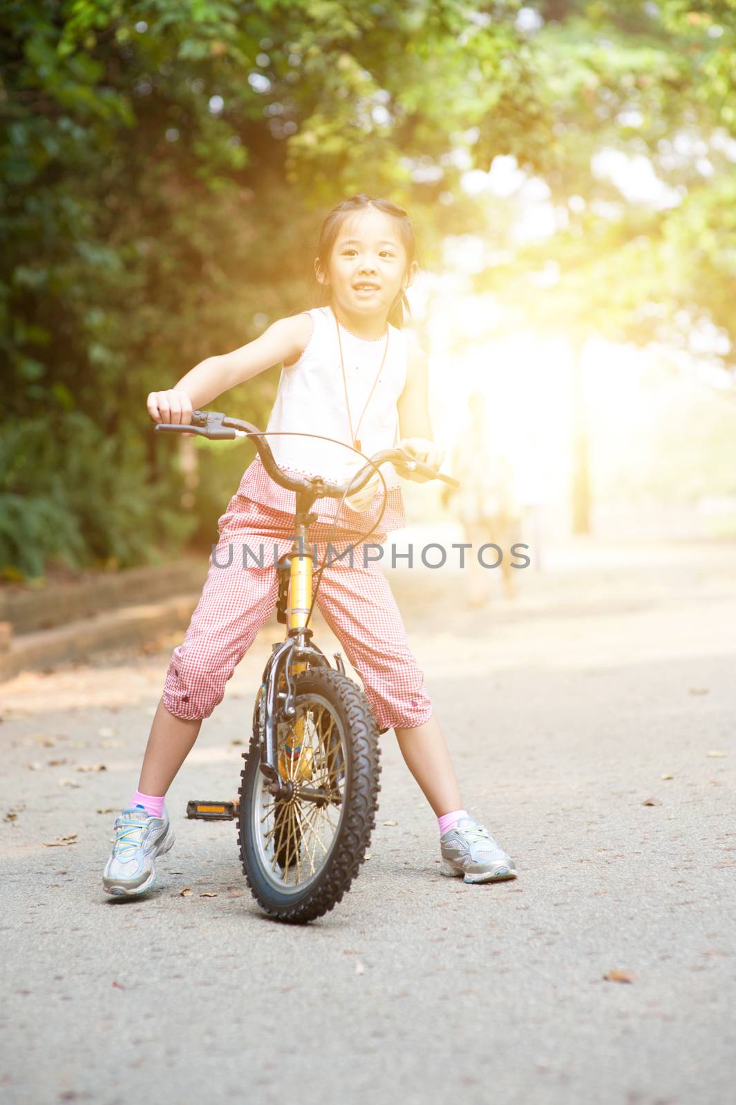 Portrait of active Asian child biking outdoors. Little girl having fun at nature park. Morning sun flare background.