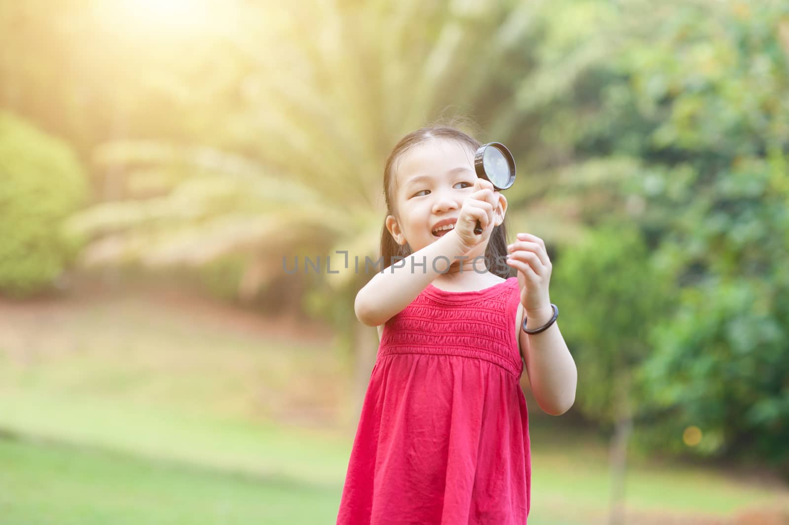 Portrait of Asian girl with magnifier glass exploring nature at park. Kid having fun outdoors. Morning sun flare background.