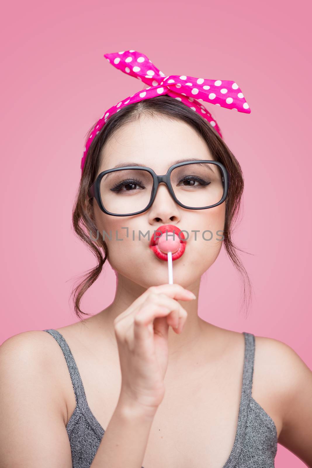 Portrait of beautiful asian woman eating heart shape lollipop, dressed and makeup in pin-up style on pink background.