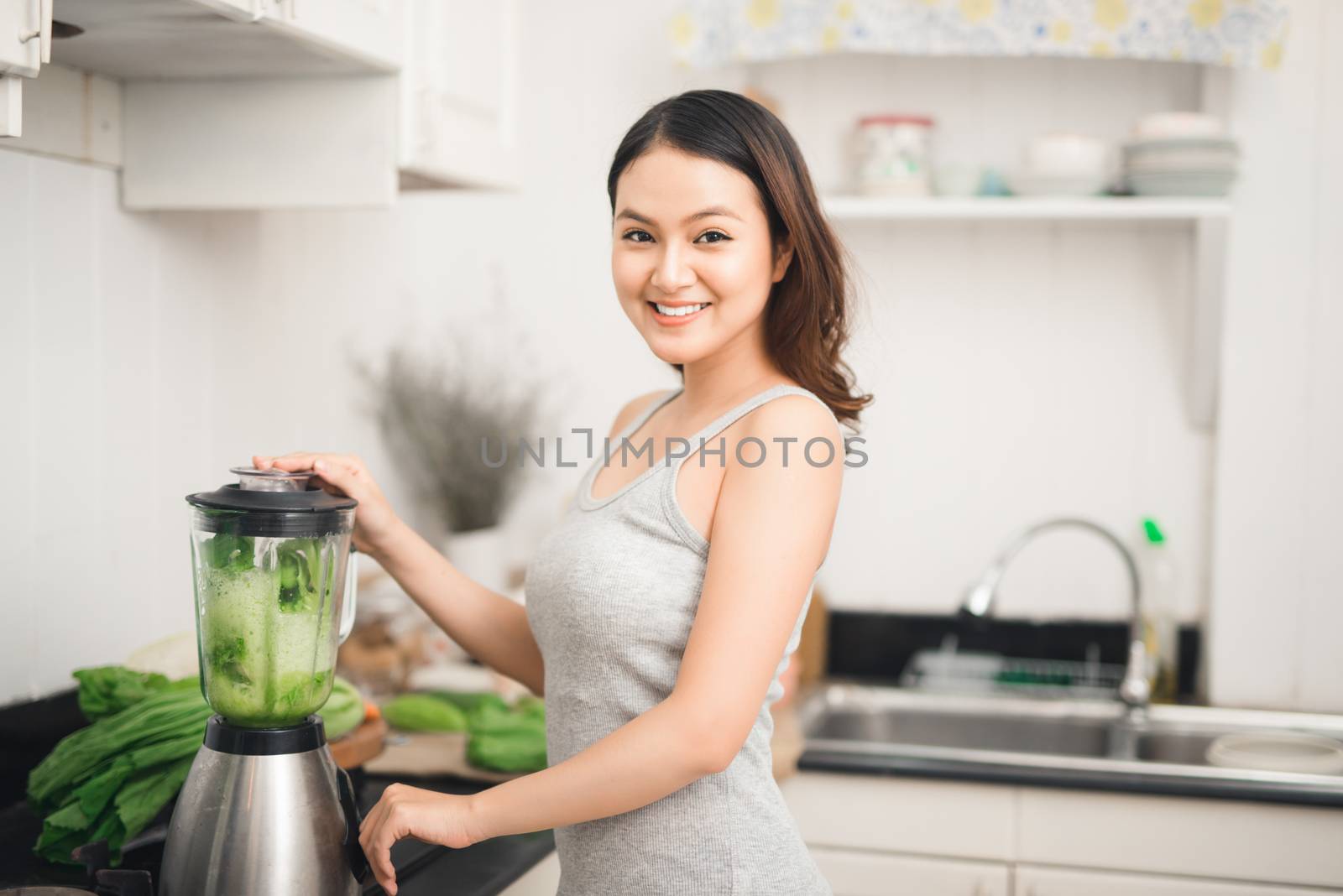 Smiling asian woman making smoothie with fresh vegetables in the blender in kitchen at home.