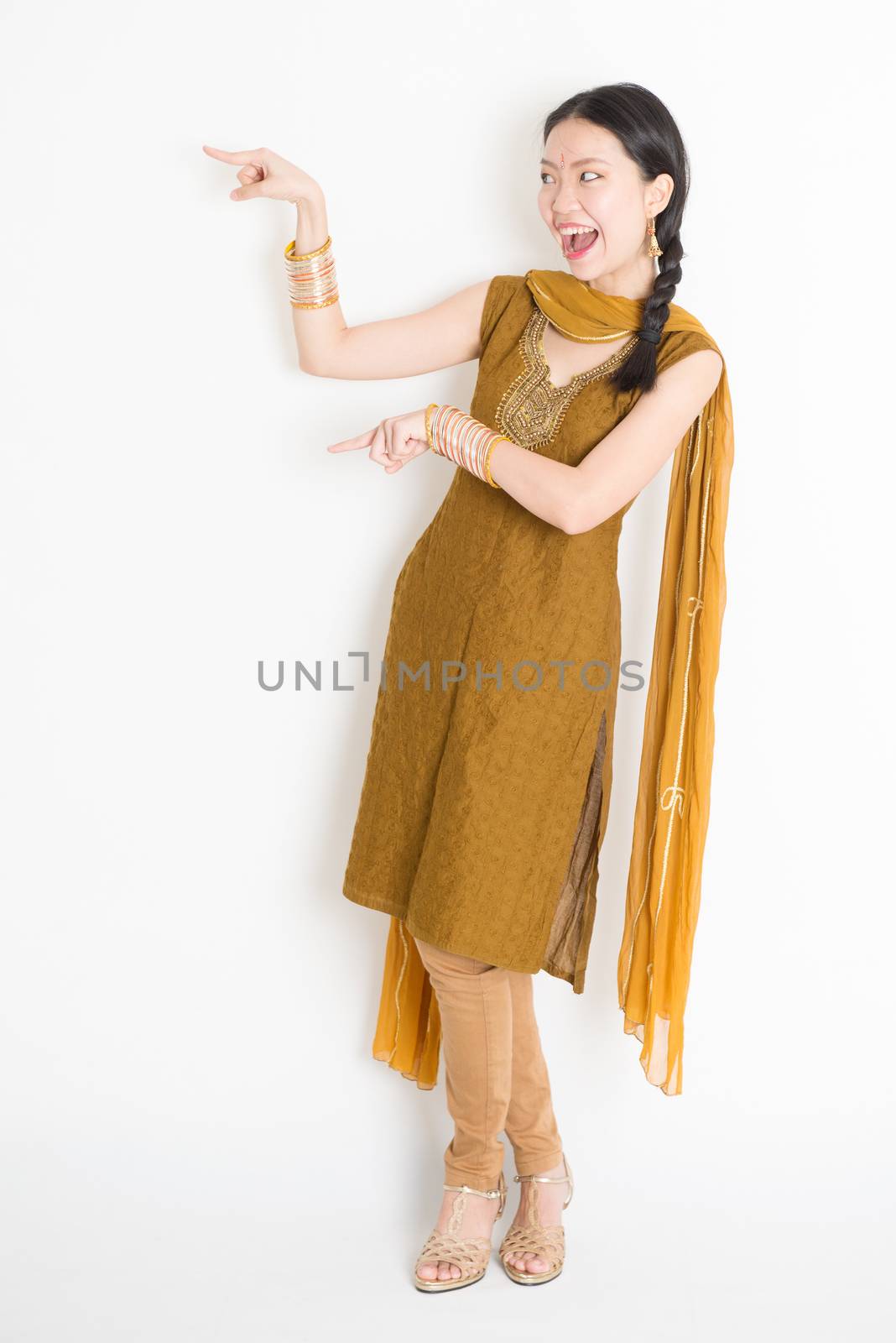 Portrait of young mixed race Indian Chinese girl in traditional punjabi dress fingers pointing at copy space, full length standing on plain white background.