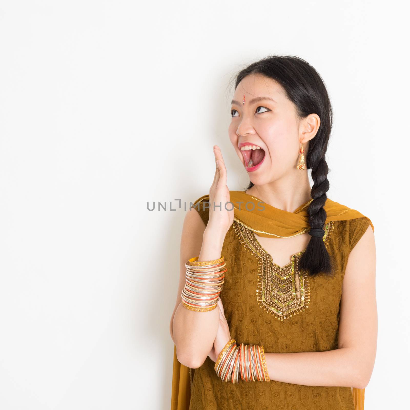 Portrait of shocked mixed race Indian Chinese woman in traditional punjabi dress opened mouth wide, surprised emotion, standing on plain white background.