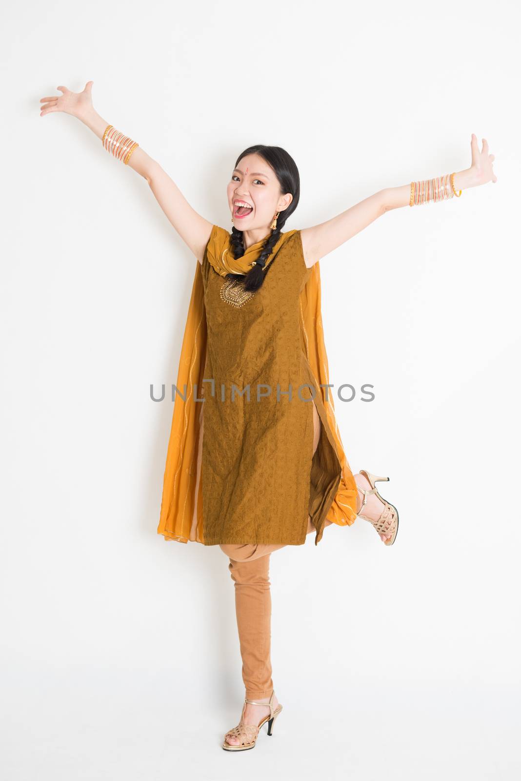 Portrait of excited mixed race Indian Chinese girl in traditional punjabi dress arms outstretched, full length standing on plain white background.