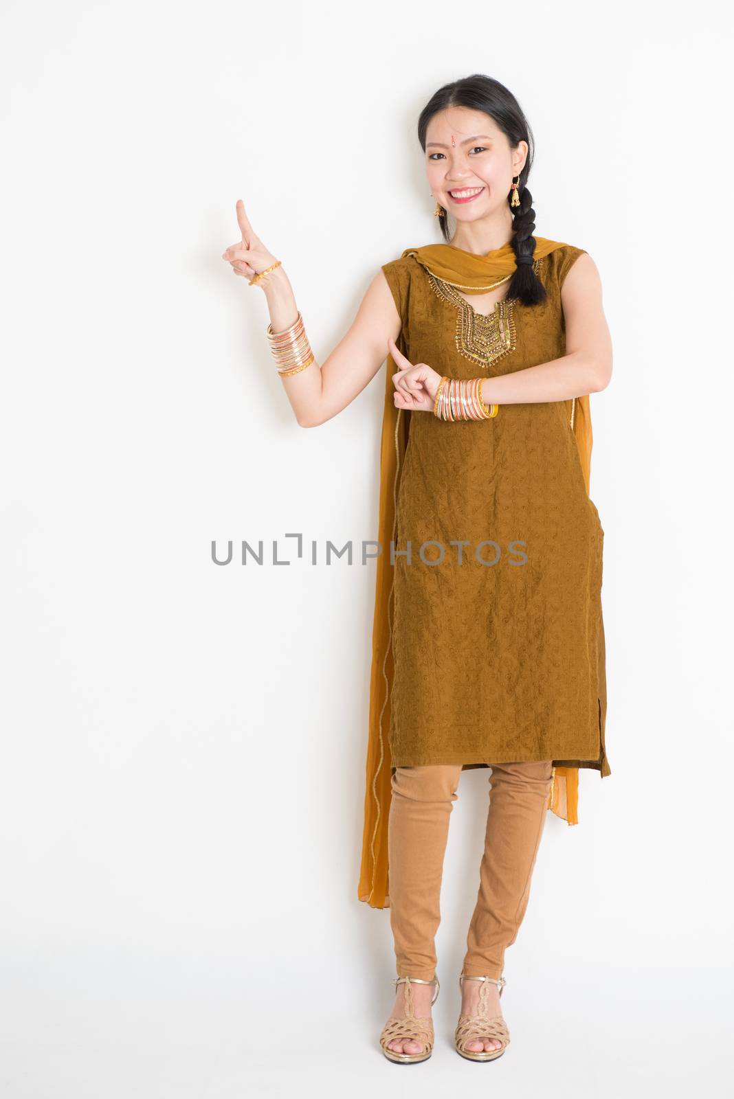 Portrait of young mixed race Indian Chinese female in traditional punjabi dress fingers pointing at copy space, full length standing on plain white background.