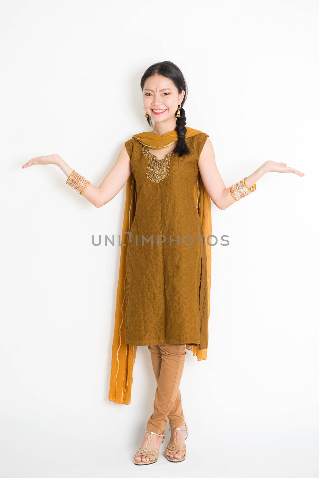 Portrait of young mixed race Indian Chinese woman in traditional punjabi dress hands showing somethings, full length standing on plain white background.