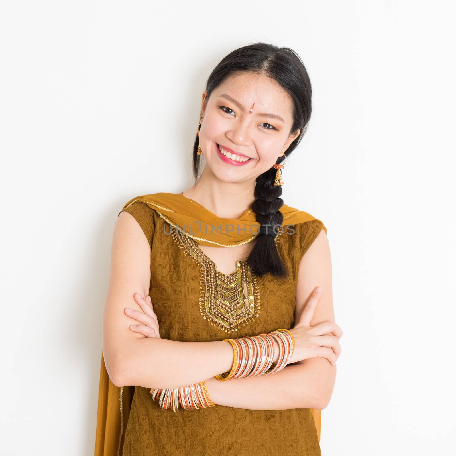 Portrait of arms crossed mixed race Indian Chinese woman in traditional Punjabi dress smiling, standing on plain white background.