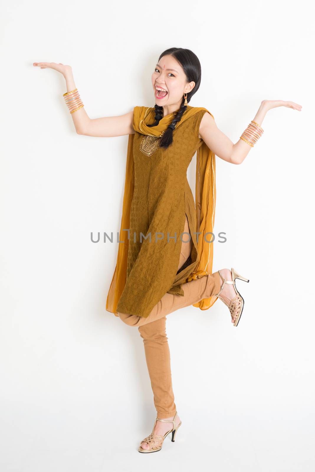 Portrait of excited mixed race Indian Chinese woman in traditional punjabi dress palms showing something, full length standing on plain white background.