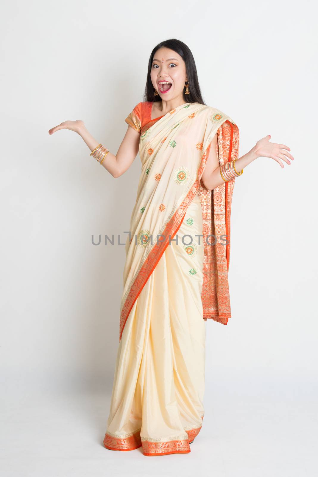 Portrait of surprised young mixed race Indian Chinese female in traditional sari dress, full length on plain background.
