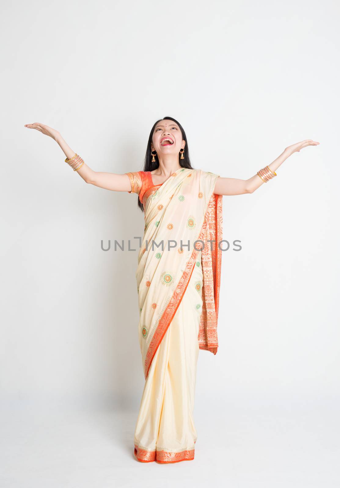 Portrait of young mixed race Indian Chinese woman in traditional sari dress hands raised looking upwards and smiling, full length on plain background.