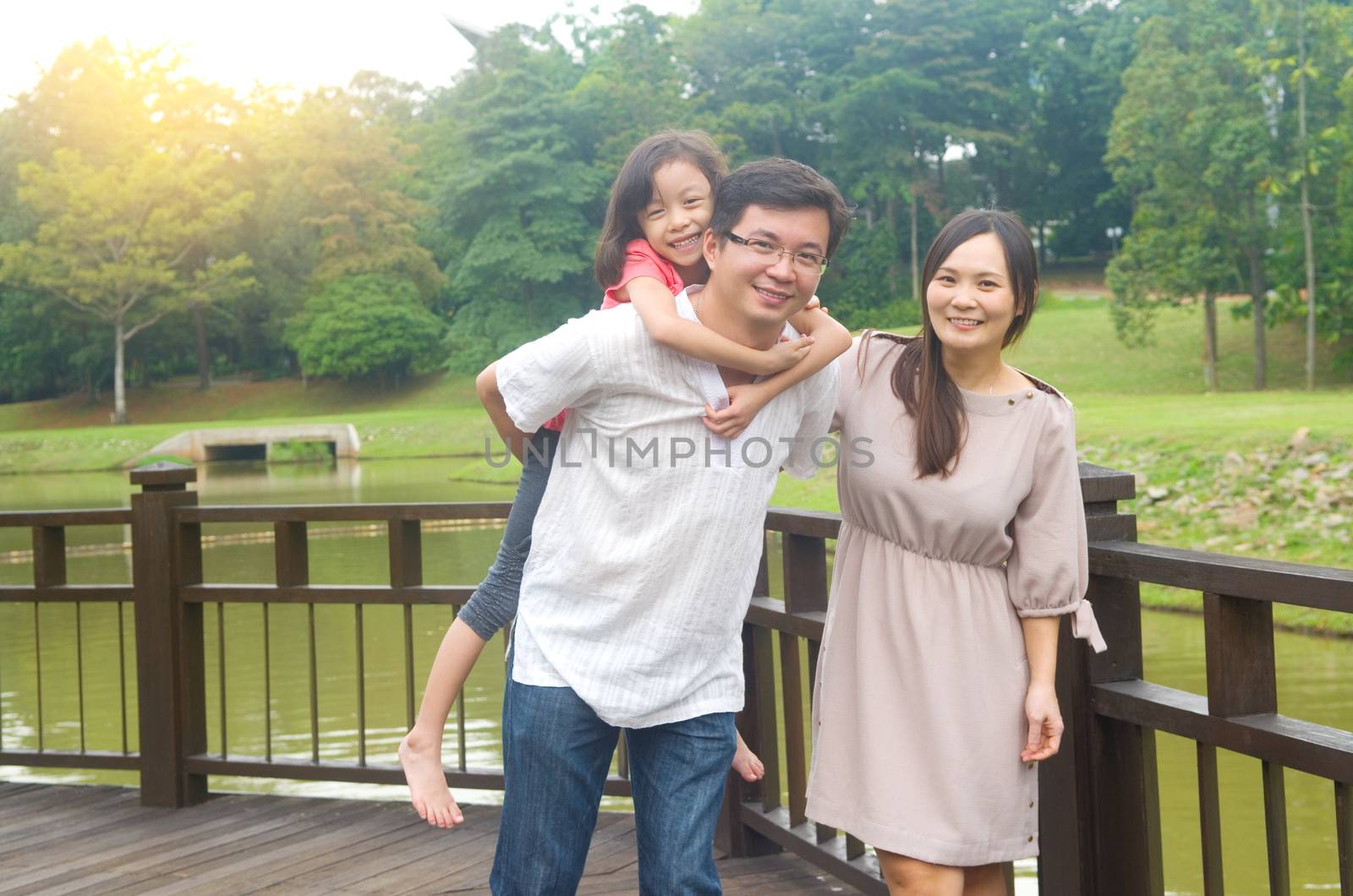 Happy Asian family outdoor. Father piggybacking his daughter walking in garden park with wife. Healthy lifestyle.