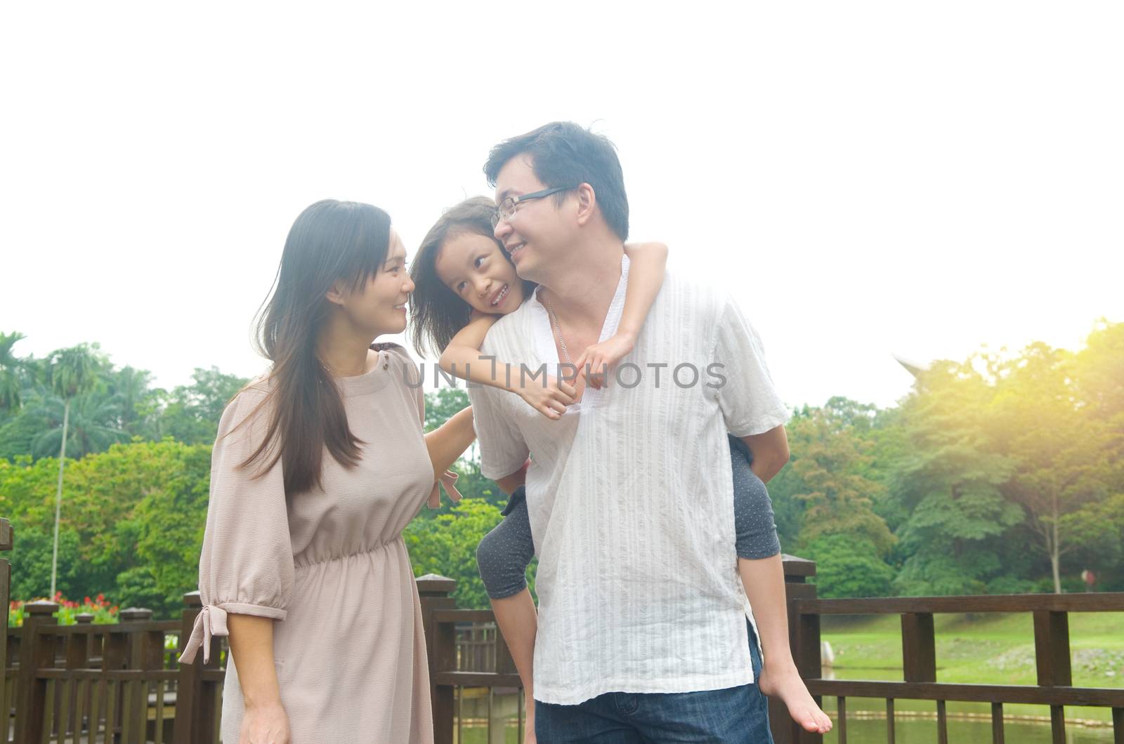 Happy Asian family outdoor. Father piggybacking his daughter walking in garden park with wife. Healthy lifestyle.