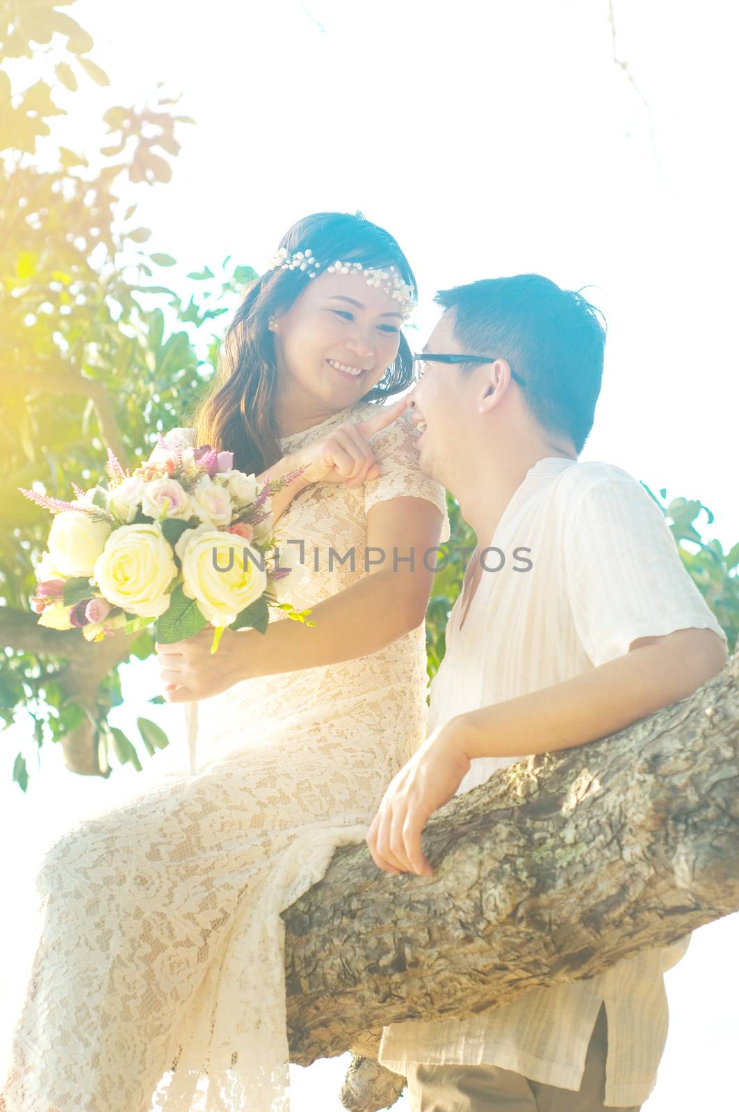 Happy asian bride sitting on the tree branch and pointed her finger on her groom's nose. Backlit shot with flare.