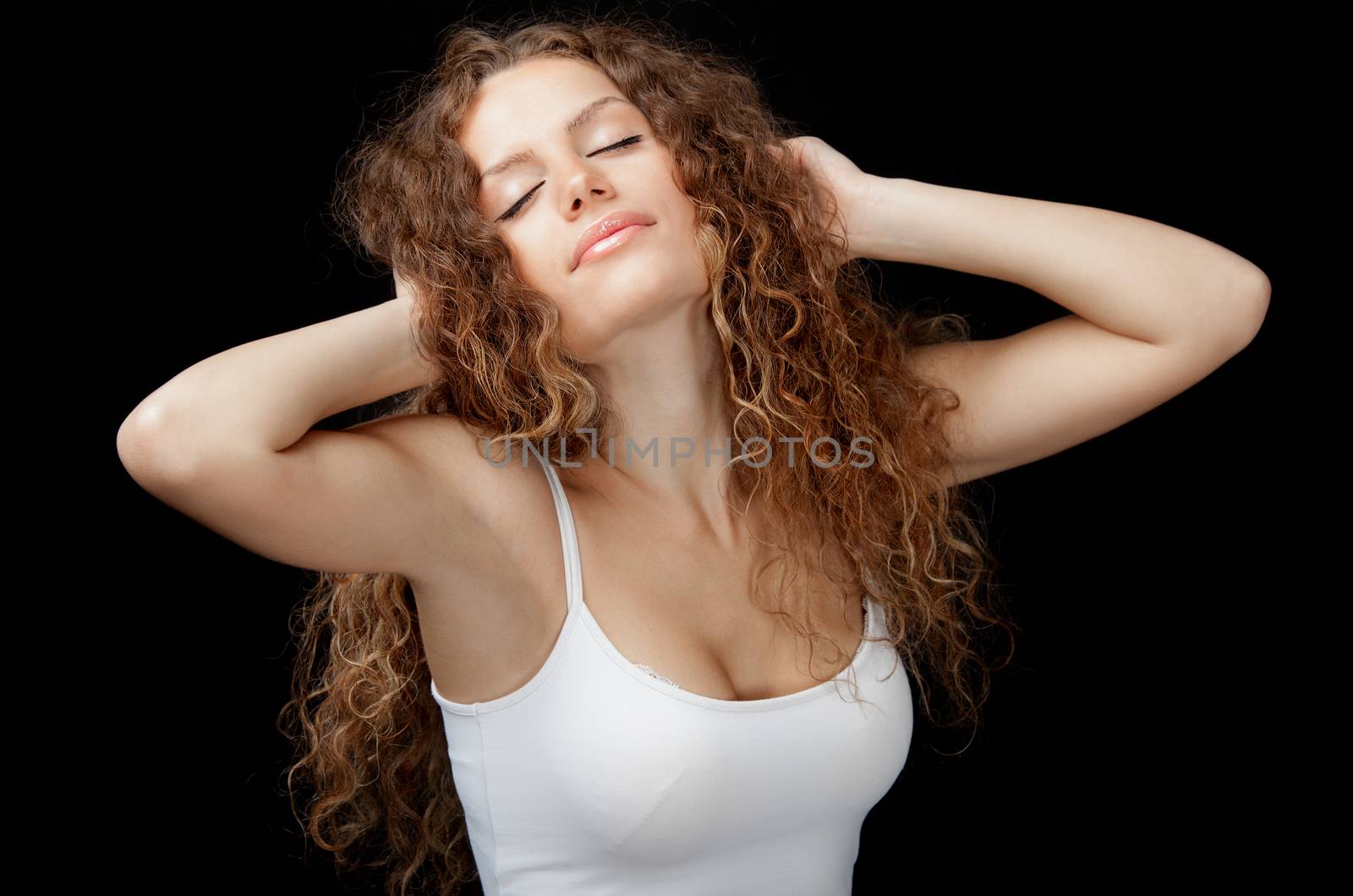 A beautiful young woman with gorgeous curly hair and white singlet stretching on black background.