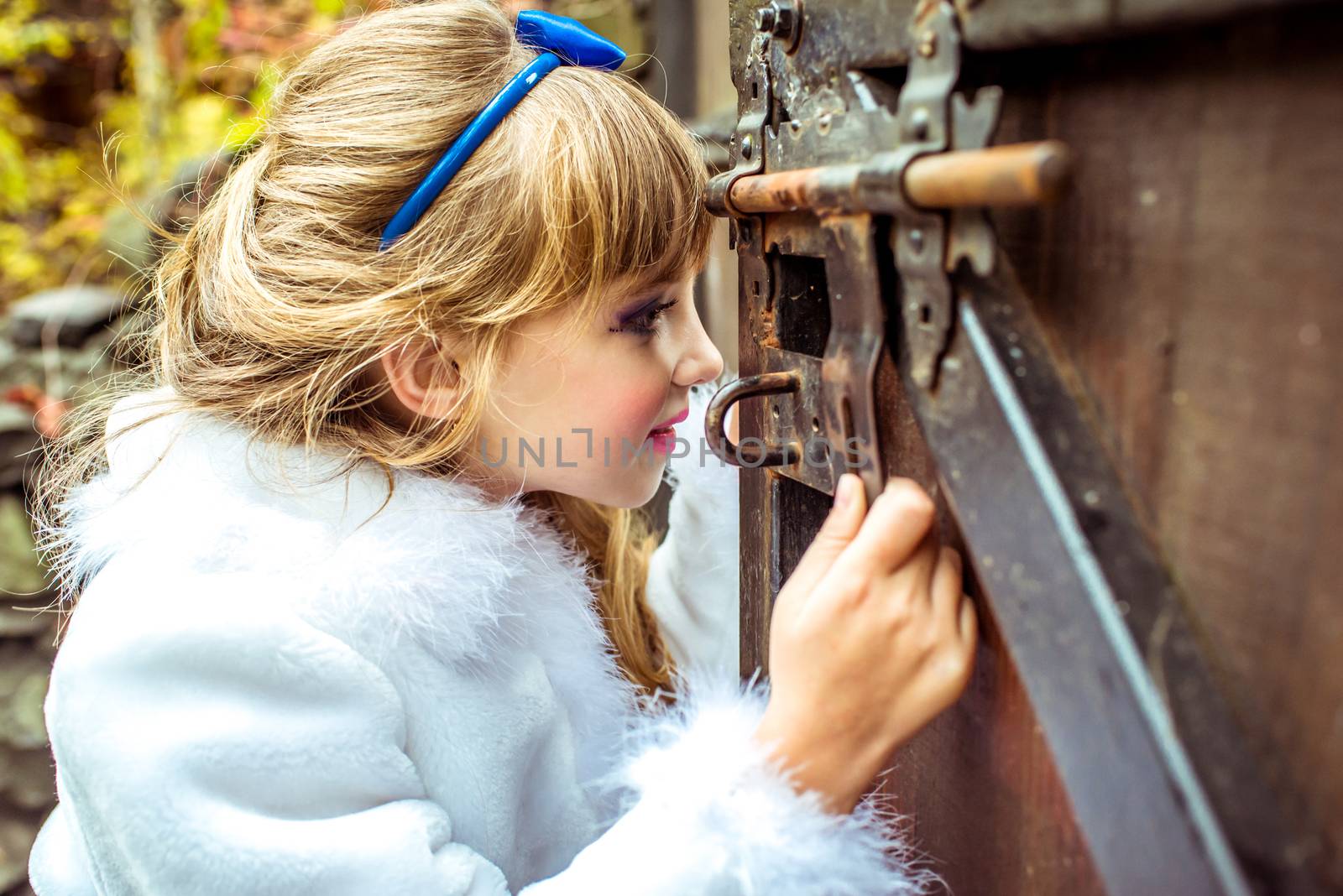 Side view of an little beautiful girl with long wavy hair in a blue silk dress in the scenery of Alice in Wonderland looking into the keyhole of the gate.