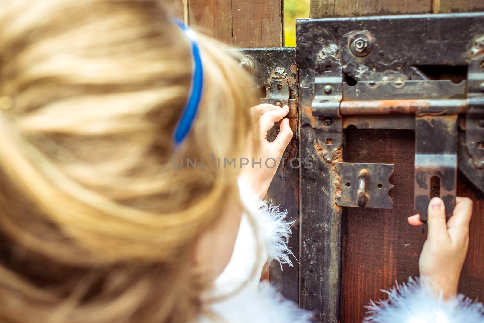 Side view of an little beautiful girl with long wavy hair in a blue silk dress in the scenery of Alice in Wonderland looking into the keyhole of the gate.