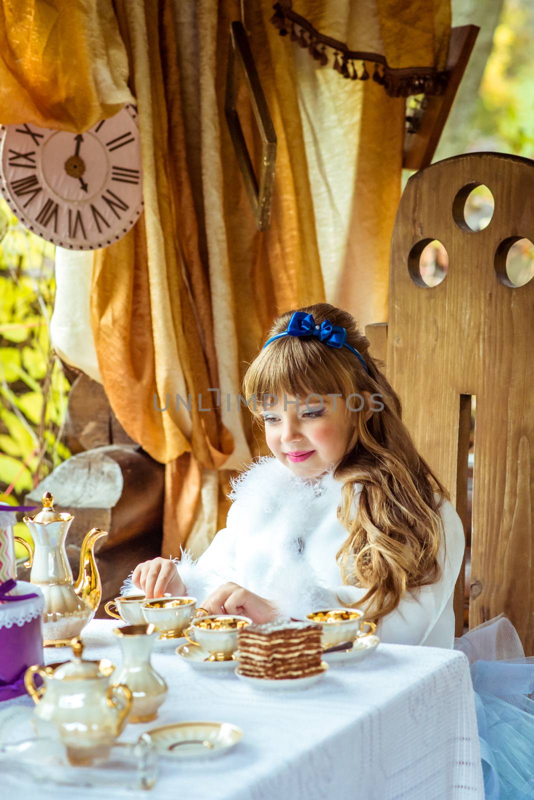 Front view of an little beautiful girl in the scenery of Alice in Wonderland holding a cup of tea at the table in the autumn park.