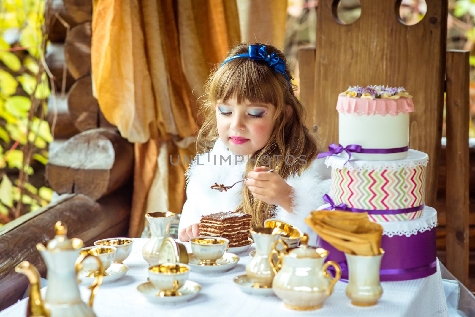 Front view of an little beautiful girl in the scenery of Alice in Wonderland holding a piece of cake on a spoon at the table in the autumn park.