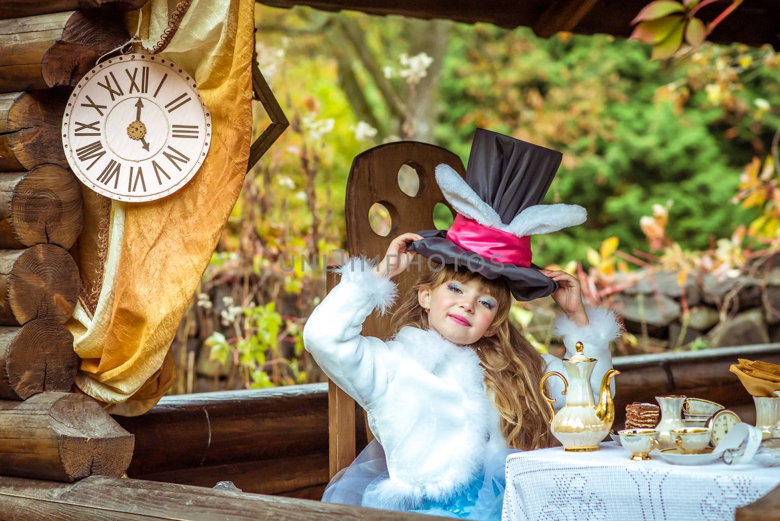 An little beautiful girl in the scenery of Alice in Wonderland holding cylinder hat with ears like a rabbit over head at the table in the garden.