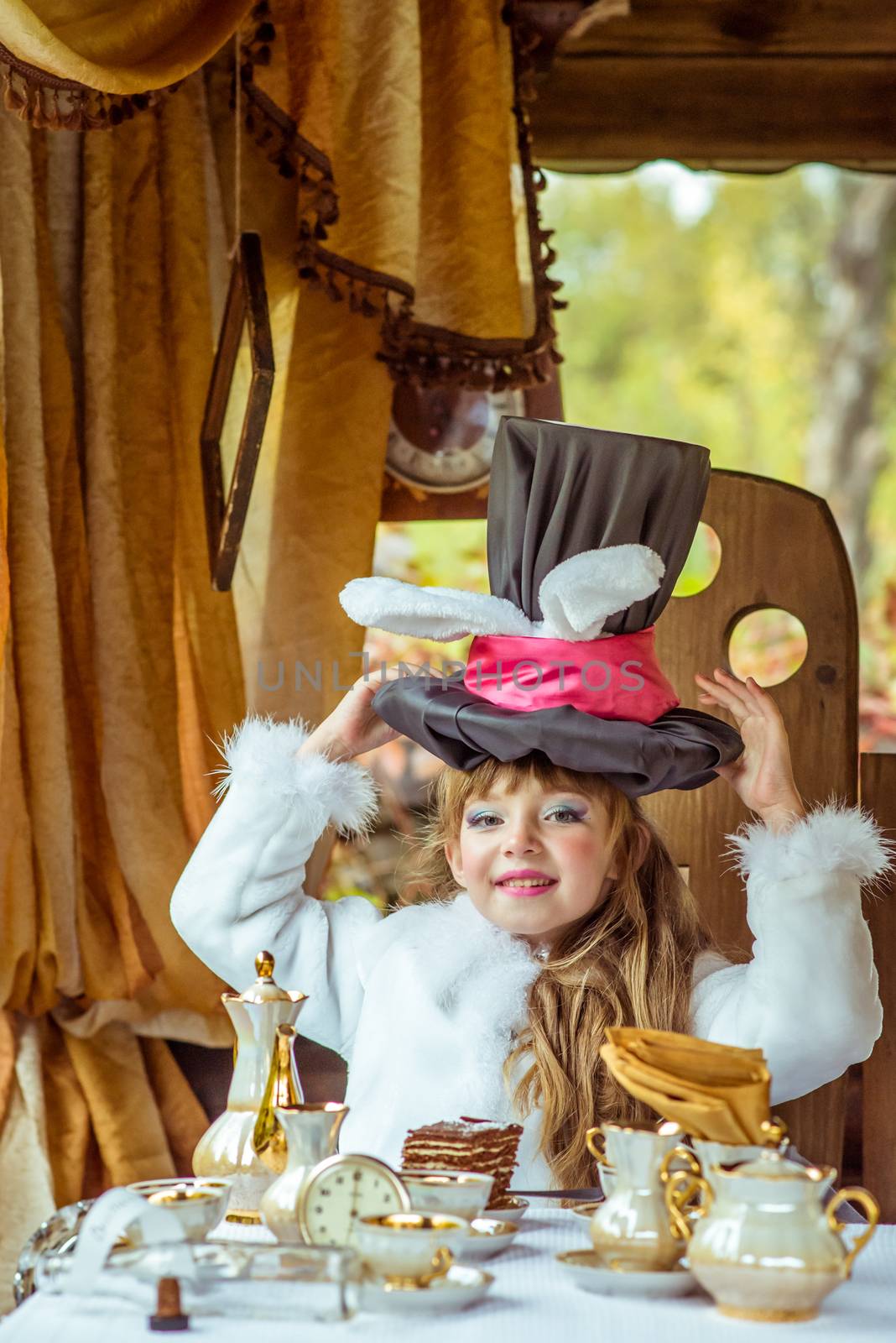 An little beautiful girl in the scenery of Alice in Wonderland holding cylinder hat with ears like a rabbit over head at the table in the garden.