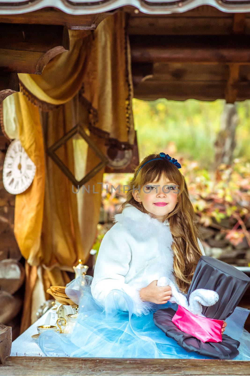 An little beautiful girl in the scenery of Alice in Wonderland holding cylinder hat with ears like a rabbit in the hands at the table in the garden.