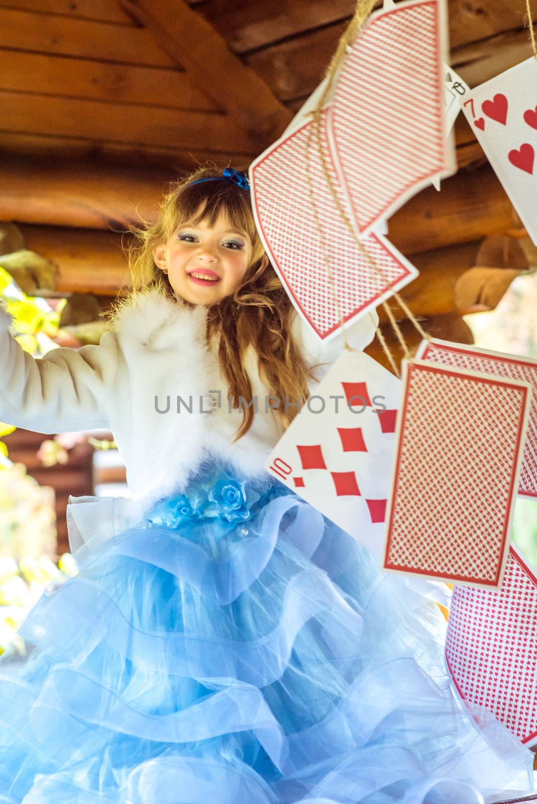 An little beautiful girl in a long blue dress in the scenery of Alice in Wonderland playing and dancing with large playing cards on the table in the garden.