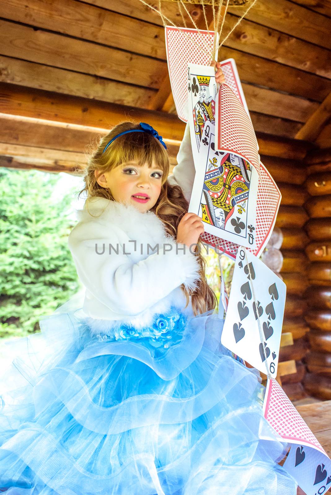 An little beautiful girl in a long blue dress in the scenery of Alice in Wonderland playing and dancing with large playing cards on the table in the garden.